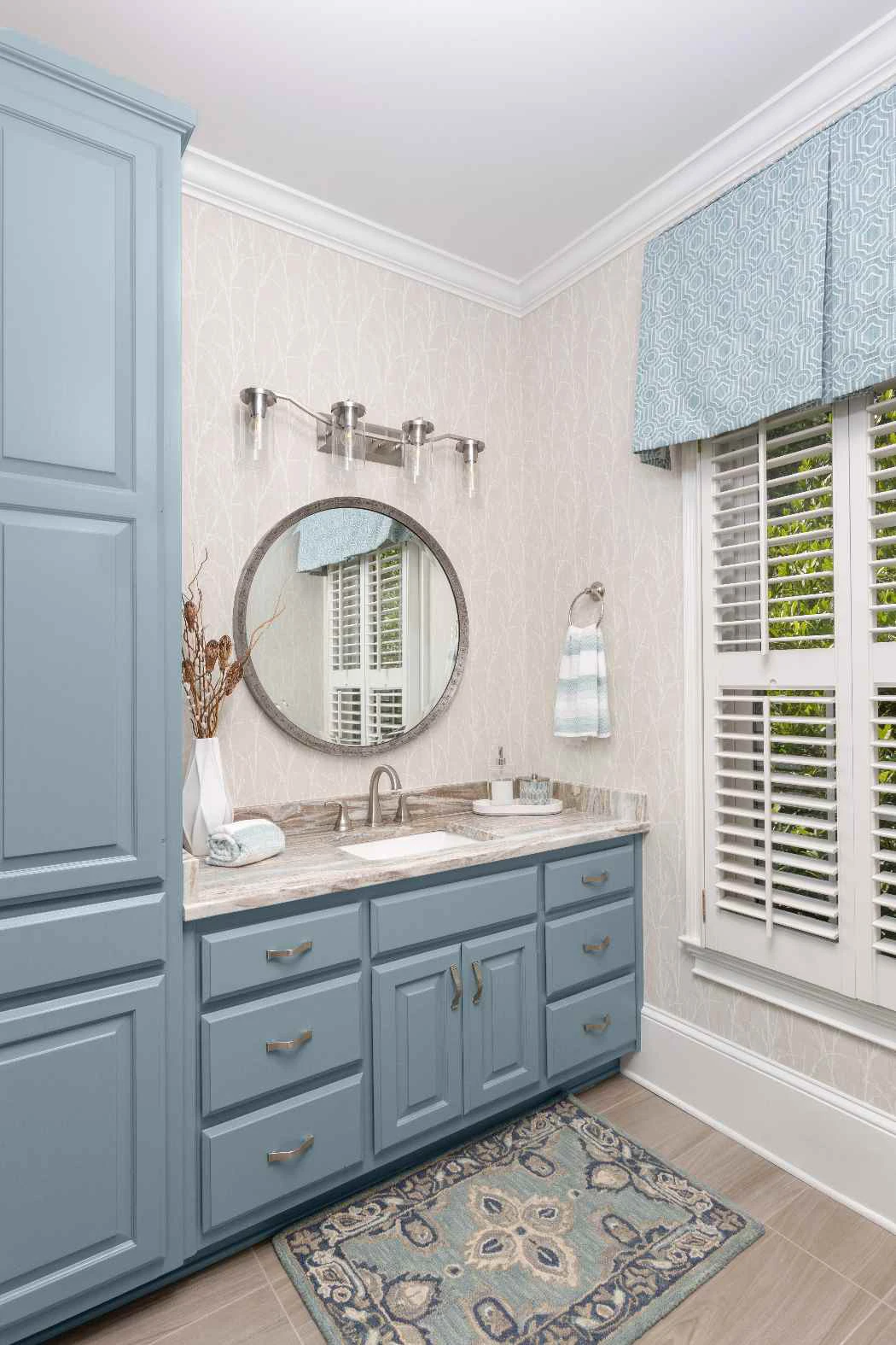 A bathroom with blue-painted cabinets, tan counters and floors, neutral wallpaper, and brushed nickel fixtures by interior designer Kristy Sherrill of Chambless Hall Design + Construction.